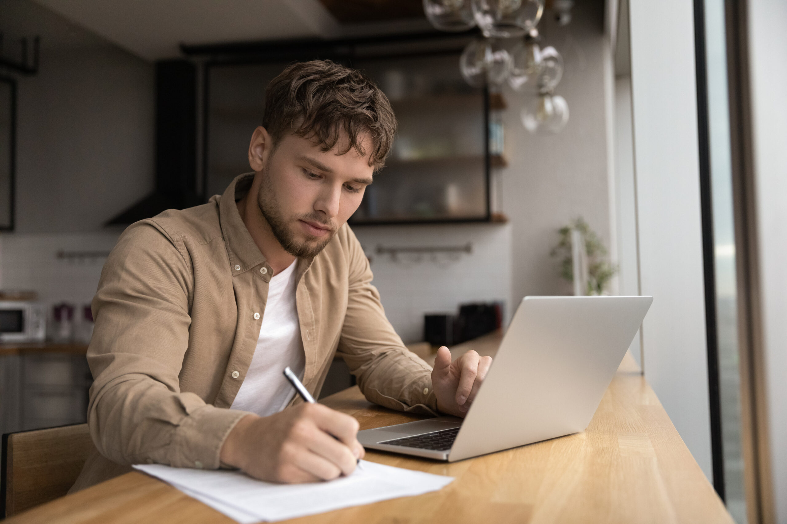 A young man writing notes on paper while working on his laptop, symbolizing the continued relevance of handwriting for learning and cognitive processing in a digital world.