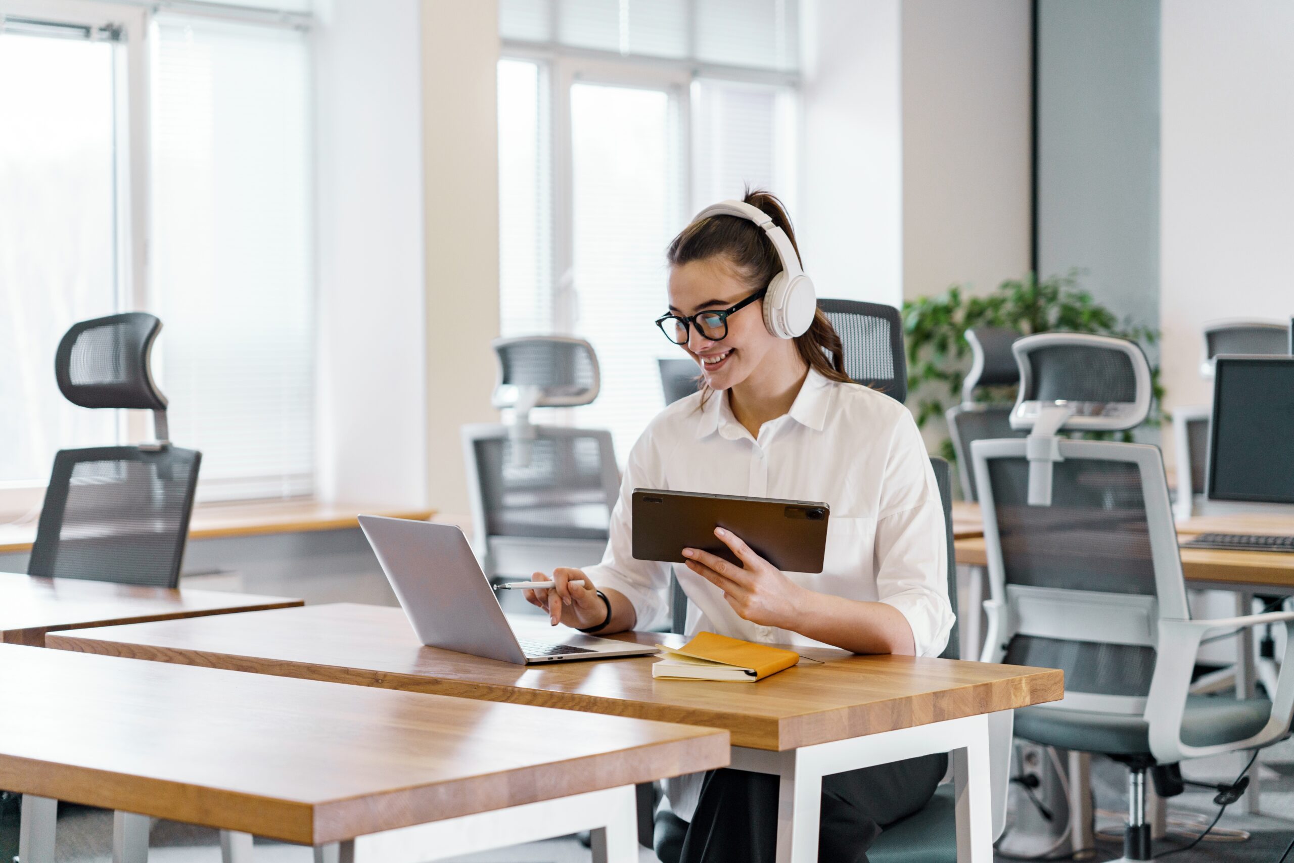 A young woman in an office wearing headphones, working on a laptop while holding a tablet, representing multitasking and its potential cognitive overload.