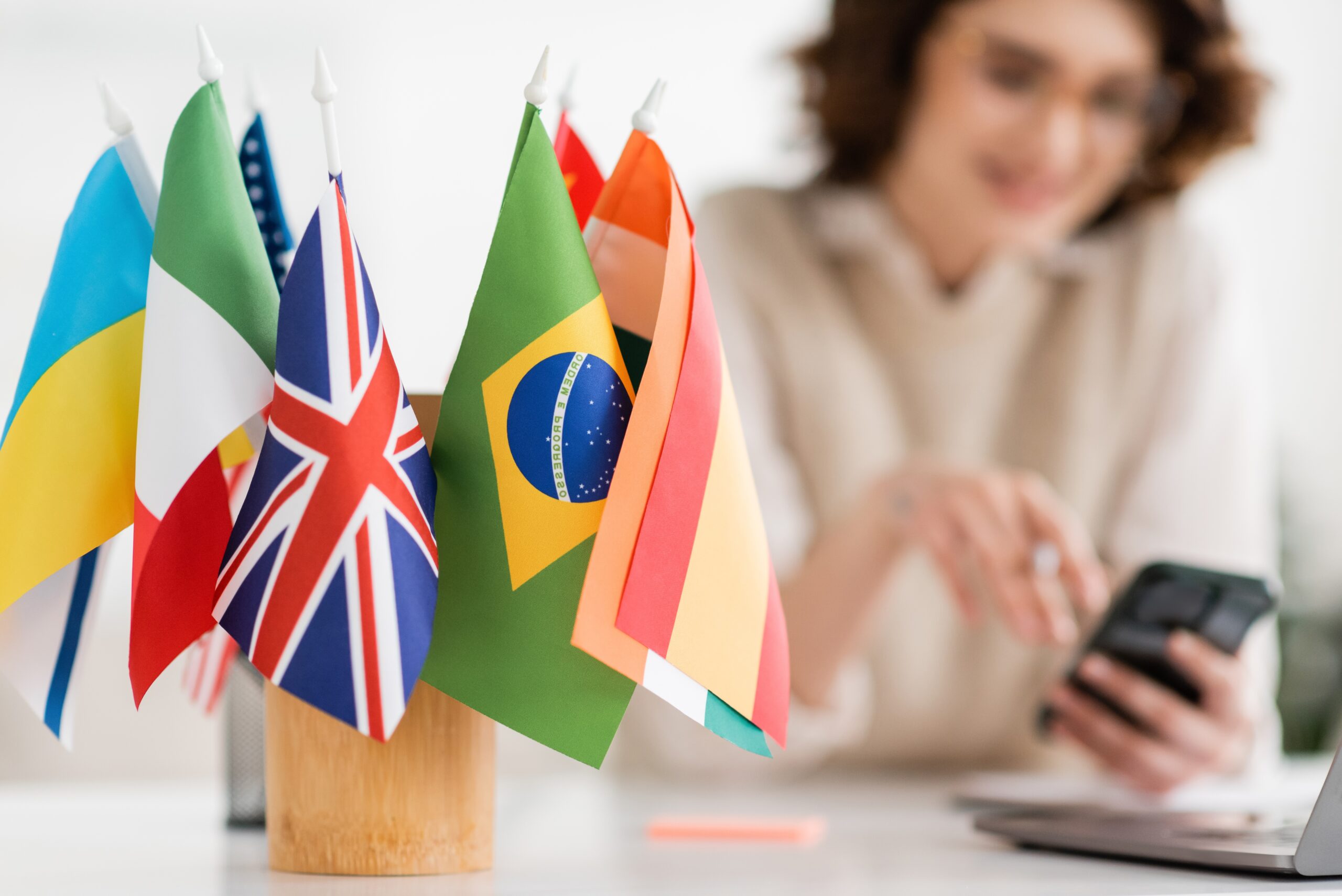 A set of international flags on a desk with a woman using a smartphone in the background, symbolizing multilingualism, cultural diversity, and cognitive benefits of learning new languages.